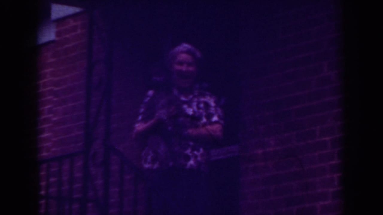 Older woman in 1962 Raleigh home-movie footage stands by a brick doorway at night while holding a small pet.