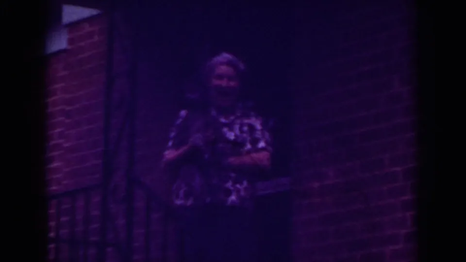 Older woman in 1962 Raleigh home-movie footage stands by a brick doorway at night while holding a small pet.