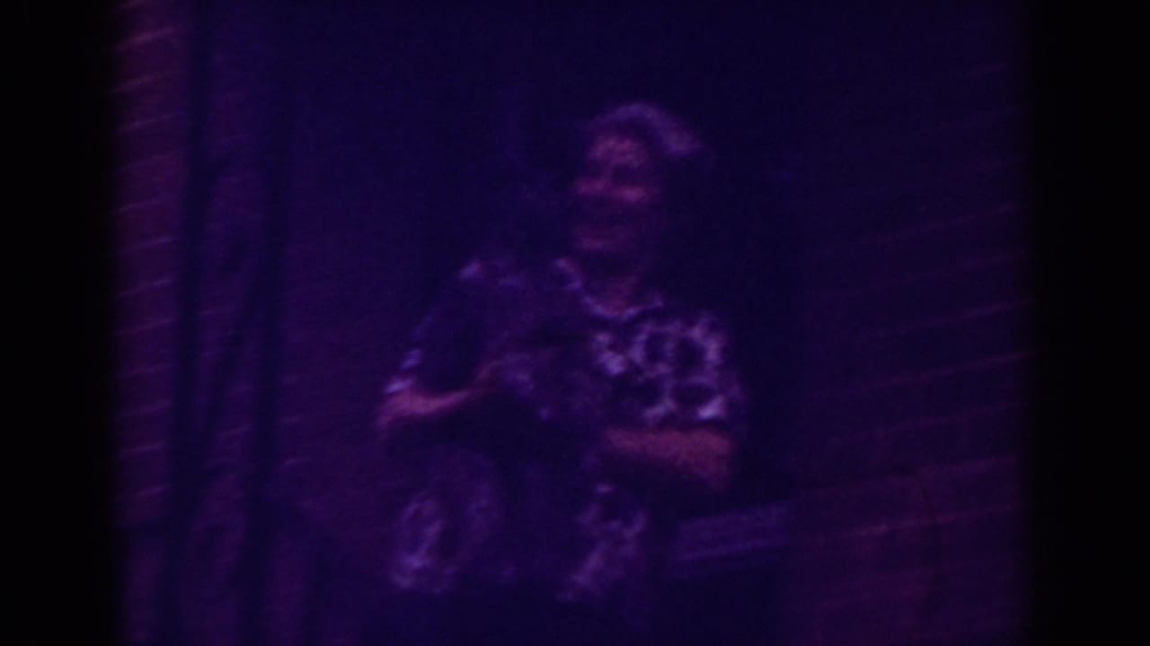 In purple-tinted 8mm footage from Raleigh, an older woman holds a small pet near a brick doorway at night.