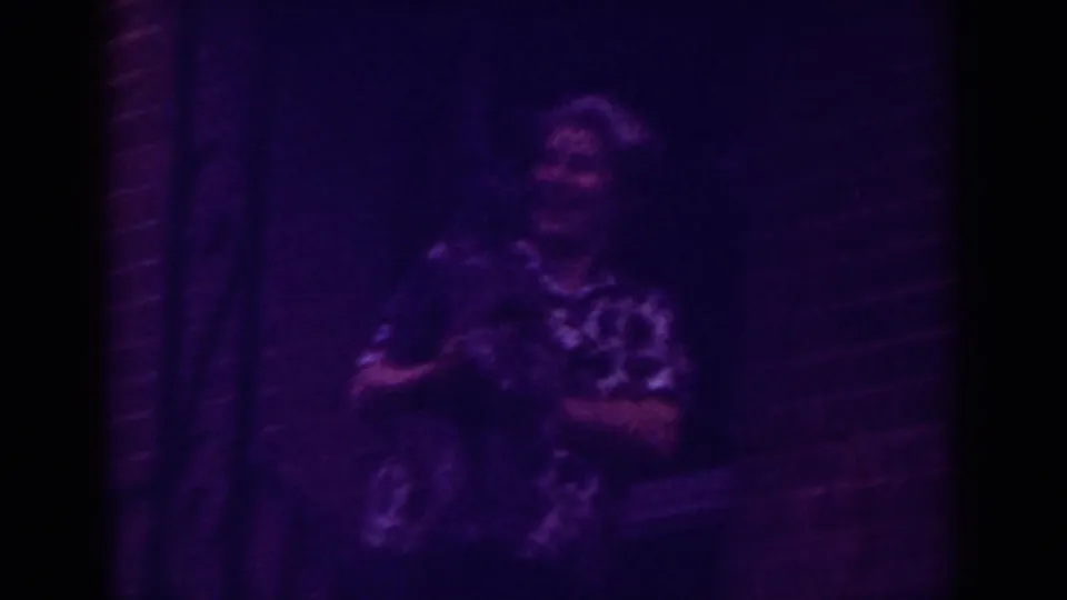 In purple-tinted 8mm footage from Raleigh, an older woman holds a small pet near a brick doorway at night.