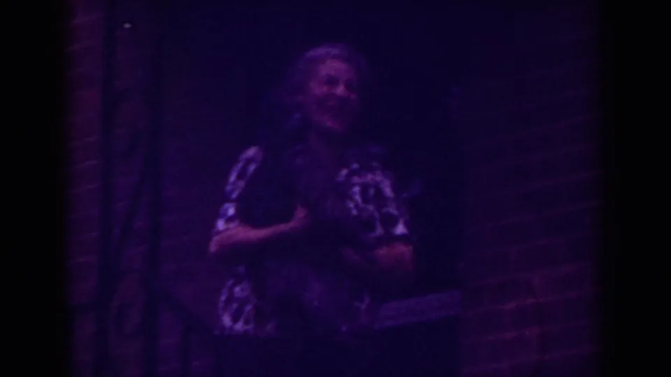 Older woman in a patterned dress looks aside while holding a small pet outside a Raleigh brick entrance.