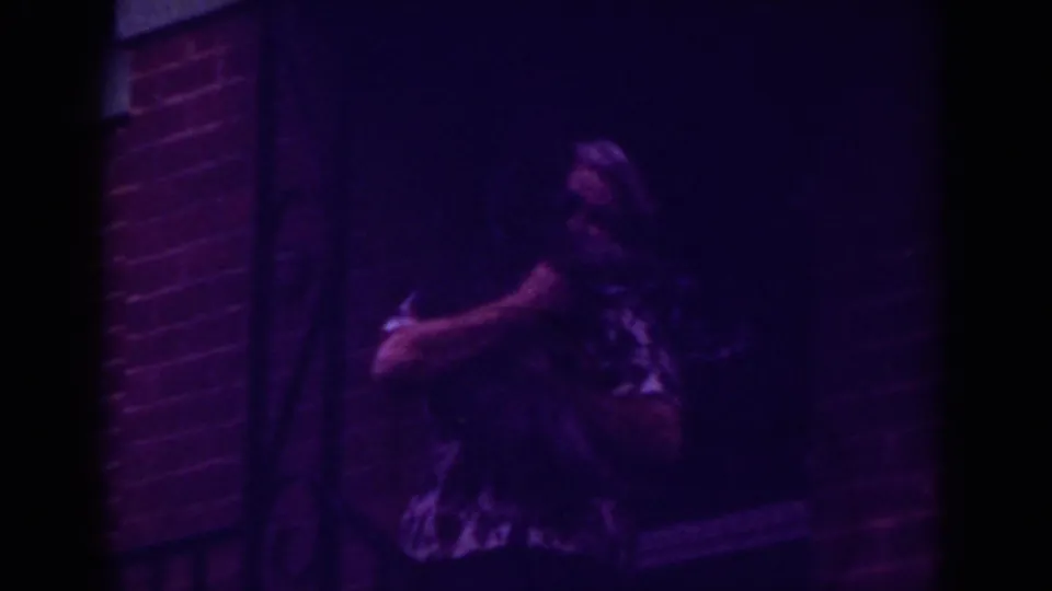 Older woman cradles a small pet beside a dark doorway and brick wall in 1962 Raleigh home-movie footage.