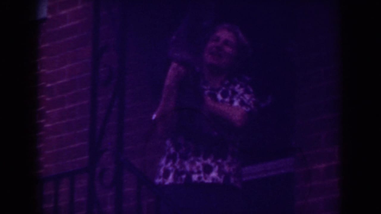 In 1962 Raleigh home-movie footage, a woman in a patterned blouse holds a small dark dog on a dim brick porch.