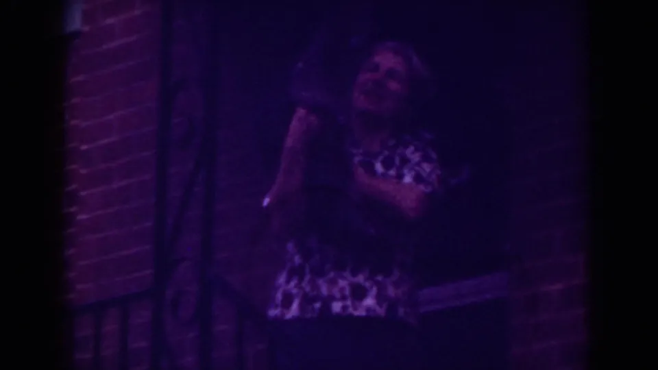 In 1962 Raleigh home-movie footage, a woman in a patterned blouse holds a small dark dog on a dim brick porch.