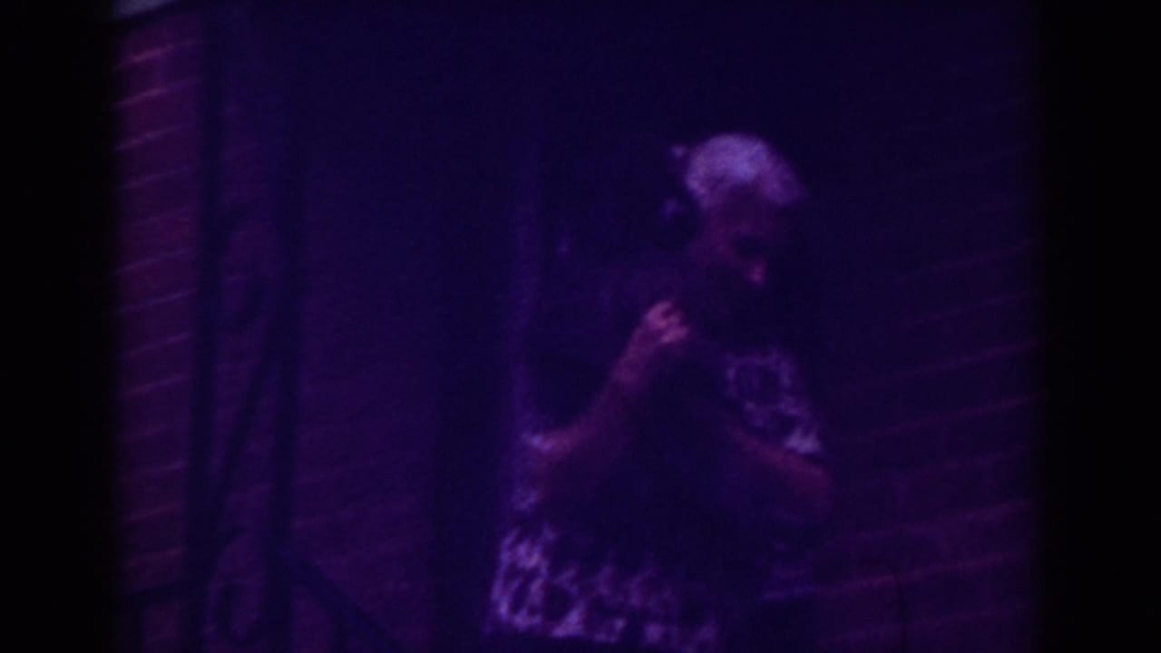 The woman turns with the small dog in her arms on a purple-tinted Raleigh porch at night.