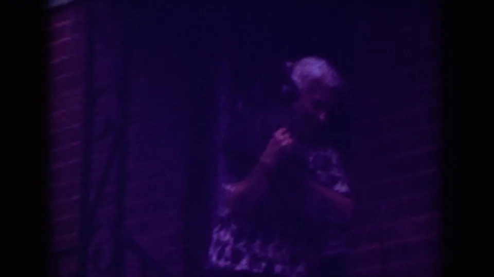 The woman turns with the small dog in her arms on a purple-tinted Raleigh porch at night.