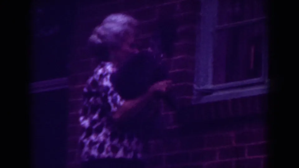 A side view shows the woman holding the dog near a brick wall and window in 1962 Raleigh footage.
