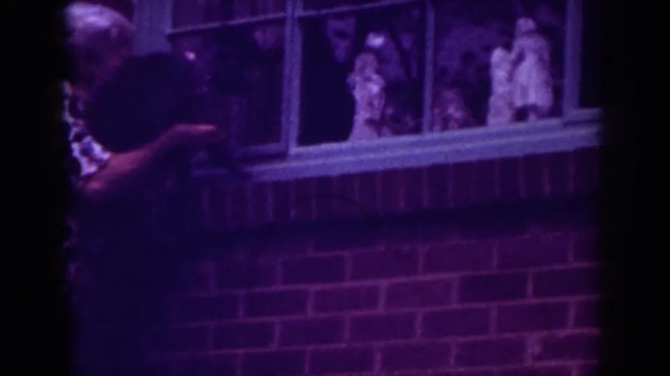 The woman holds the small dog beside porch windows, with small decorative figures visible beyond the glass in Raleigh footage.