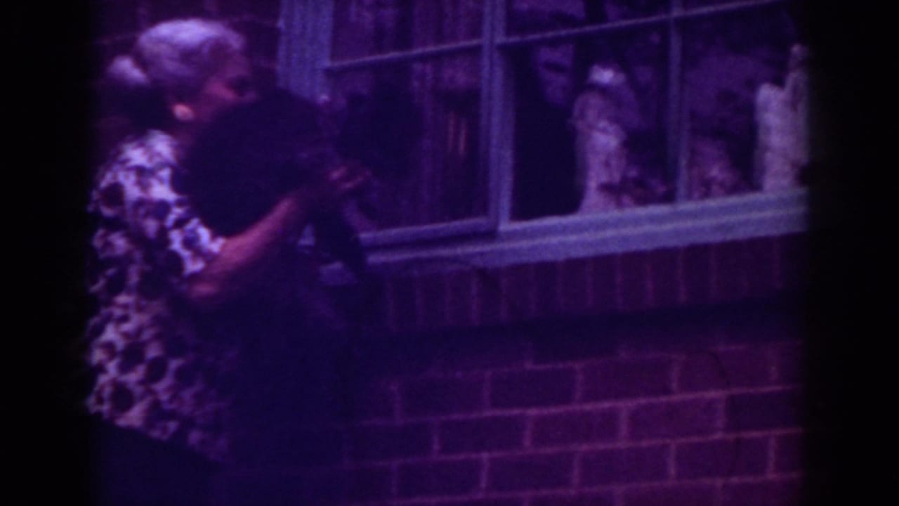 In metadata-supported 1962 Raleigh footage, a woman holds a black dog up toward a brick house window with figures visible behind glass.