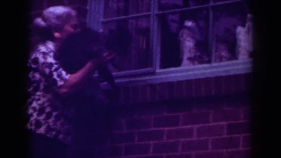 In metadata-supported 1962 Raleigh footage, a woman holds a black dog up toward a brick house window with figures visible behind glass.