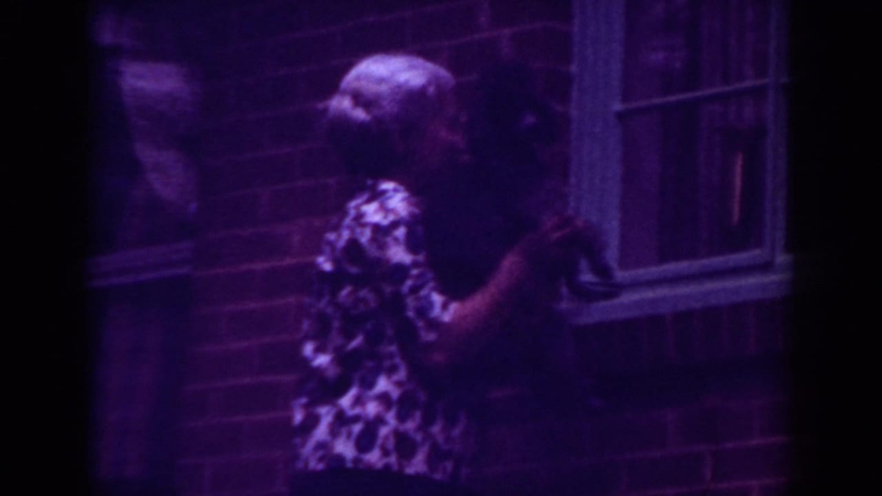 Woman stands beside a brick wall holding a black dog near a window.