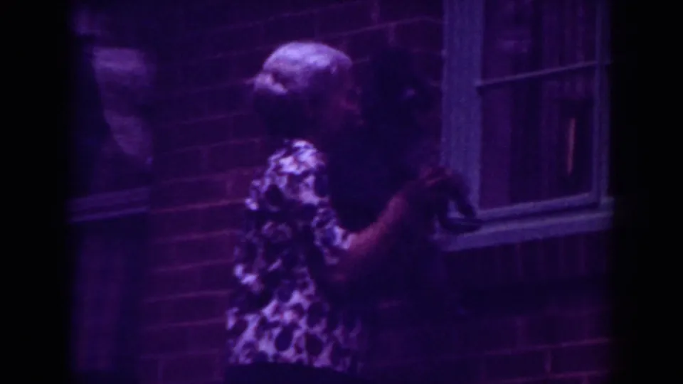 A woman in a patterned blouse holds a black dog beside a brick exterior and window in purple-tinted 8mm home-movie footage.