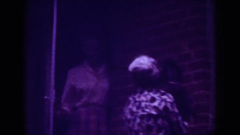 Two adult women appear outside a brick house, one in a light shirt and plaid skirt and the other seen from behind in a patterned blouse.