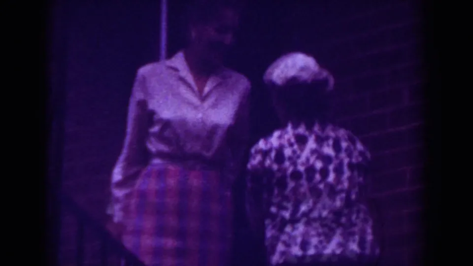 Two women stand near a brick wall in 1962 Raleigh home-movie footage, with one facing camera and another seen from behind.
