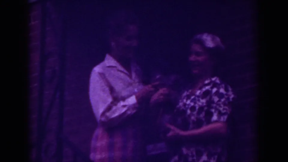 The women face one another at the Raleigh doorway, with one holding or passing a small dark item in soft purple-tinted 8mm footage.