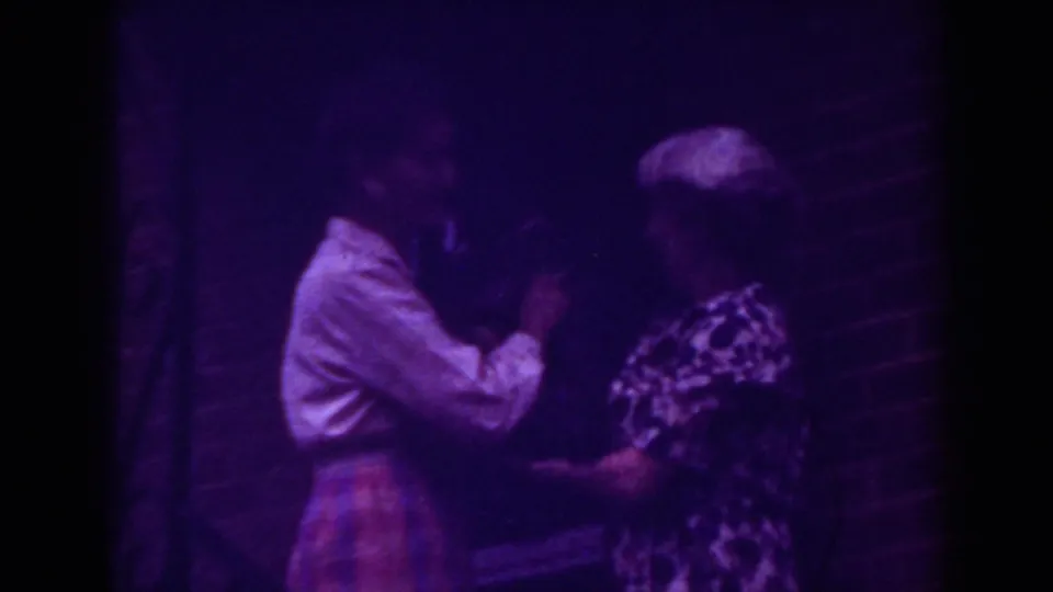 One woman leans in toward the other near the brick doorway, suggesting a close greeting or reaction during the 1962 home-movie moment.