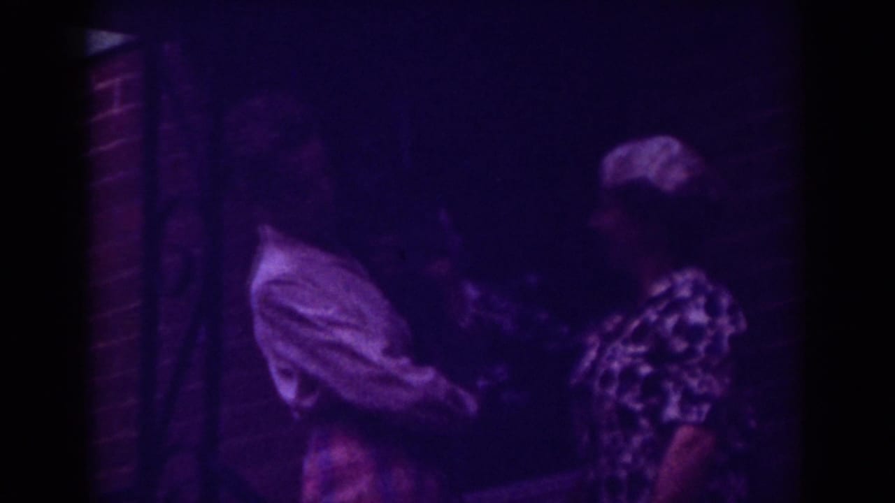 1962 Raleigh home-movie frame showing two women close together near a brick wall, one in a light collared shirt and one in a patterned dress.