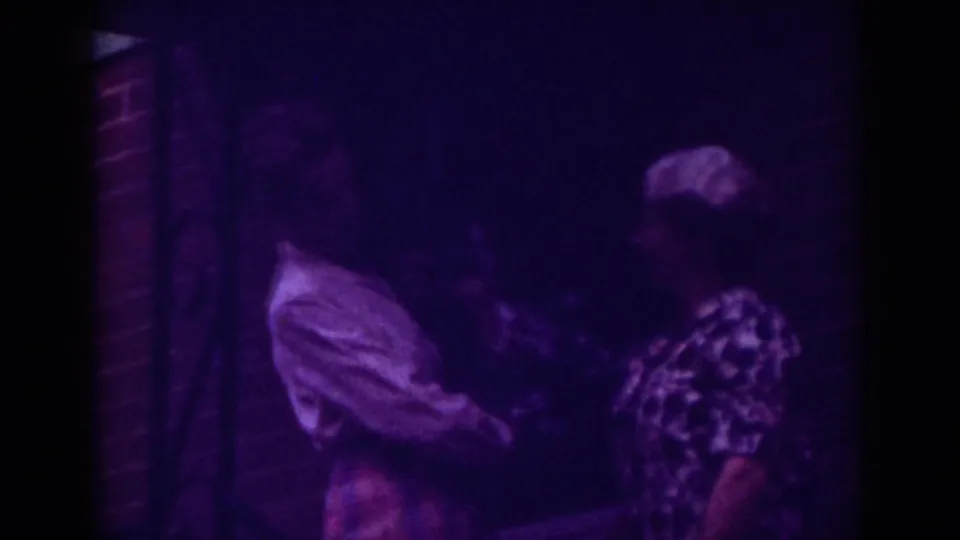 1962 Raleigh home-movie frame showing two women close together near a brick wall, one in a light collared shirt and one in a patterned dress.
