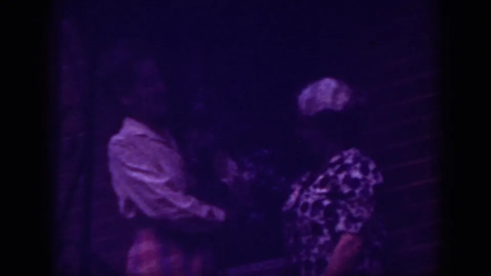 1962 Raleigh home-movie still showing two women close together near brickwork, with heavy grain, low light, and purple color shift.