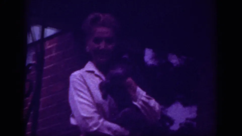 Raleigh 1962 8mm frame with a woman in a light collared shirt embracing another figure in a dark, purple-tinted outdoor setting.