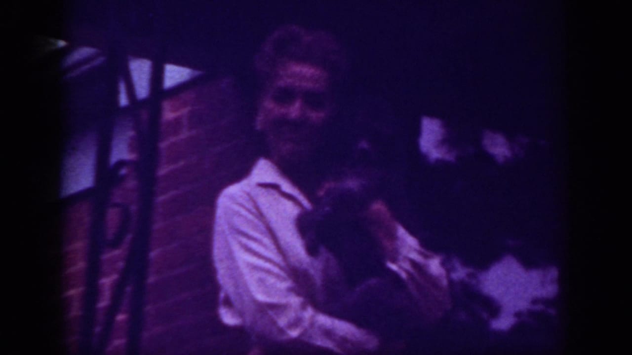 1962 Raleigh home-movie frame showing a woman in a white shirt holding a dark-furred pet near a brick wall at night.