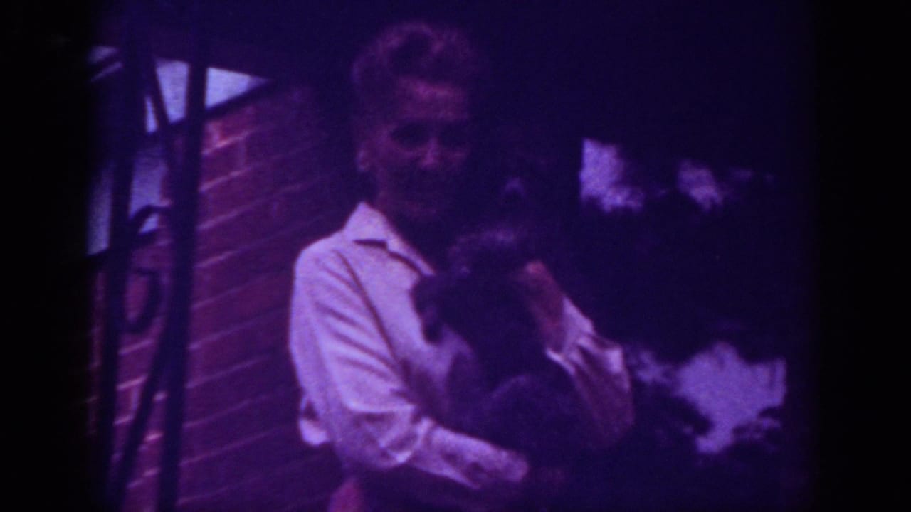 1962 Raleigh home-movie frame showing a woman outside a brick house holding a dark animal in low light.