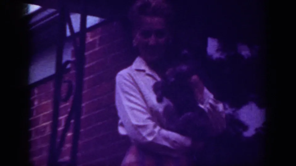Home-movie frame from 1962 Raleigh showing the woman facing camera with a dark animal in her arms outside the house.