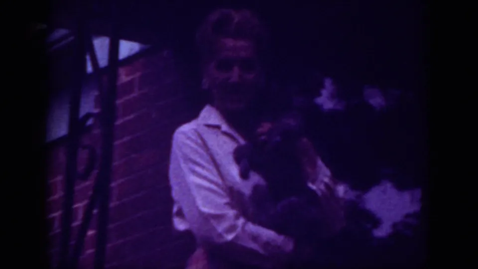 1962 Raleigh home-movie still of a woman holding a dark animal beside a brick house under dim nighttime lighting.