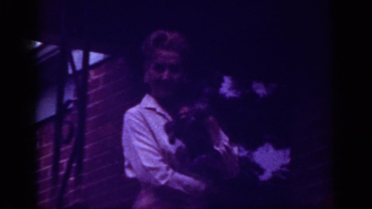 1962 Raleigh home-movie frame showing a woman on a brick porch holding a small dark dog under a purple color cast.