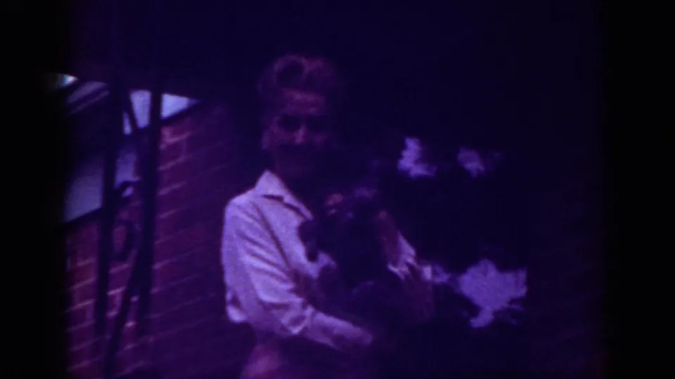1962 Raleigh home-movie frame showing a woman on a brick porch holding a small dark dog under a purple color cast.