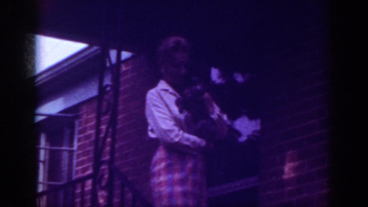 Woman stands on a brick porch holding a small dog near a railing and window.