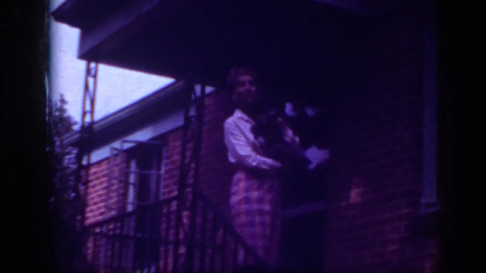 1962 Raleigh home-movie frame showing a woman holding a dog on a brick porch beneath an overhang.