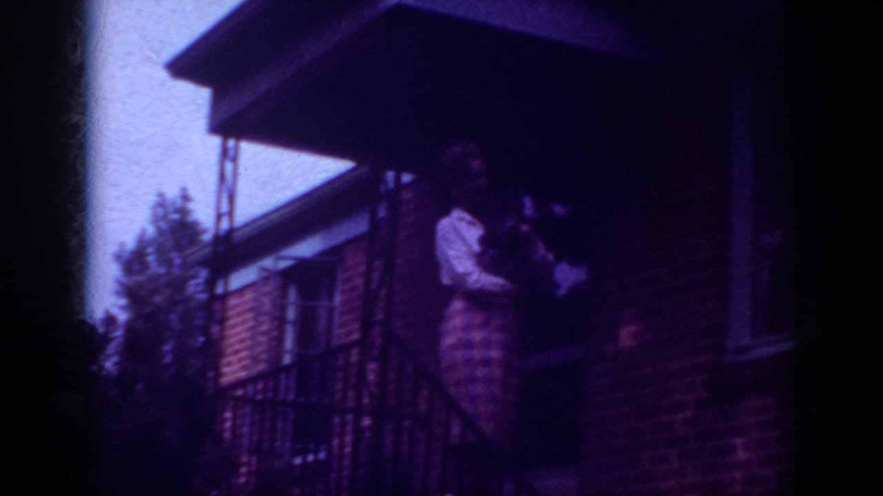 Adult woman stands on a brick porch holding a small dog near a metal railing.
