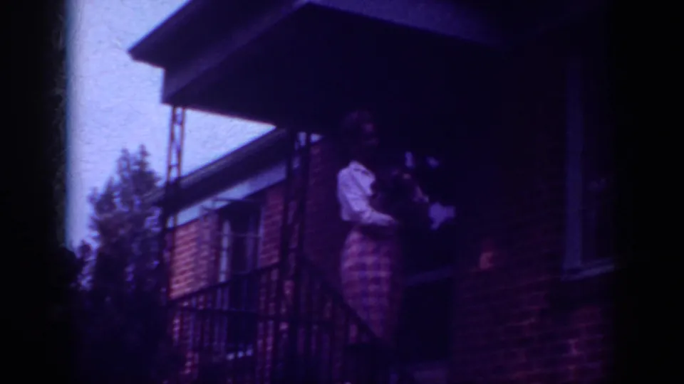 In 1962 Raleigh home-movie footage, an adult woman in a light blouse and plaid skirt stands on a brick porch holding a small dog.