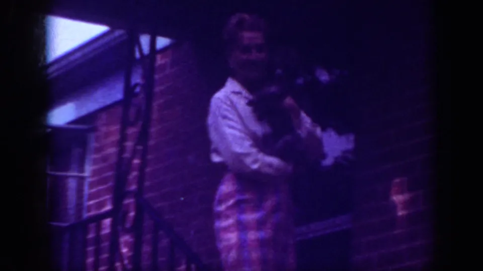 A closer 8mm frame shows the woman smiling while holding the small dog on the brick porch in Raleigh, with strong purple color shift.