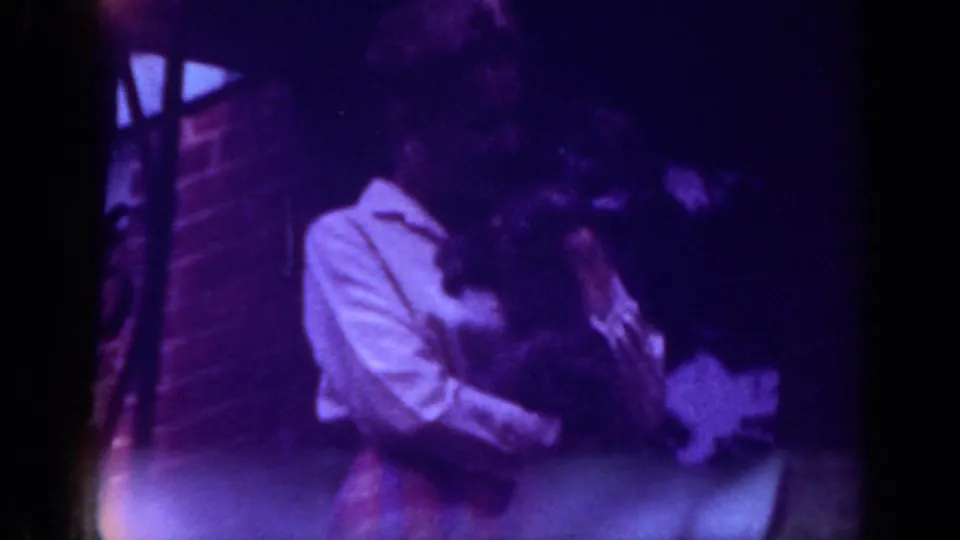 The woman leans in to kiss the small dog she is holding on the Raleigh porch, framed tightly in soft, purple-shifted 8mm footage.
