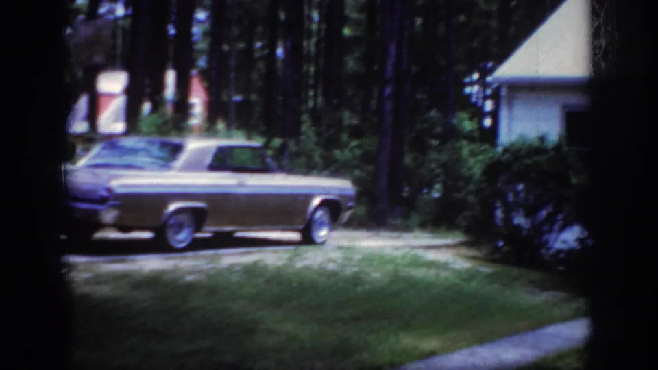 Home-movie frame of a two-tone sedan outside a modest house amid trees and lawn in 1962 Raleigh.