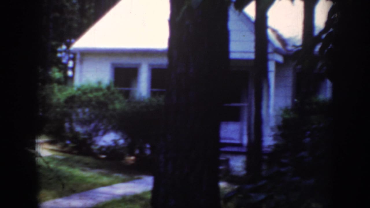 1962 Raleigh home-movie frame showing a white house partly obscured by trees, shrubs, lawn, and a walkway.