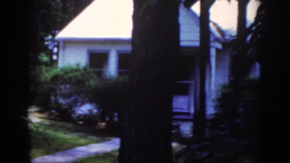 1962 Raleigh home-movie frame showing a white house partly obscured by trees, shrubs, lawn, and a walkway.