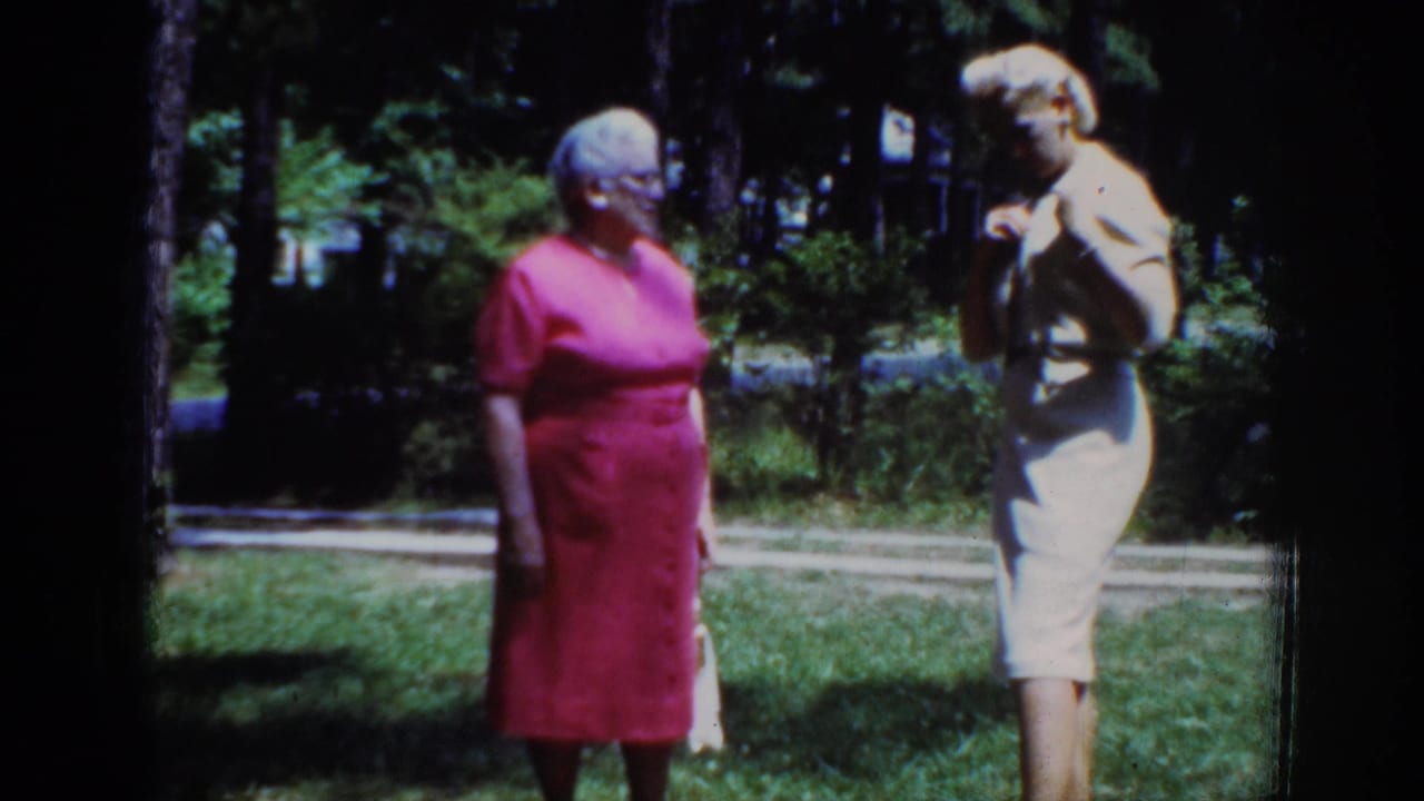 1962 Raleigh home-movie frame showing two adult women conversing outdoors, with grass, trees, and strong sunlight behind them.