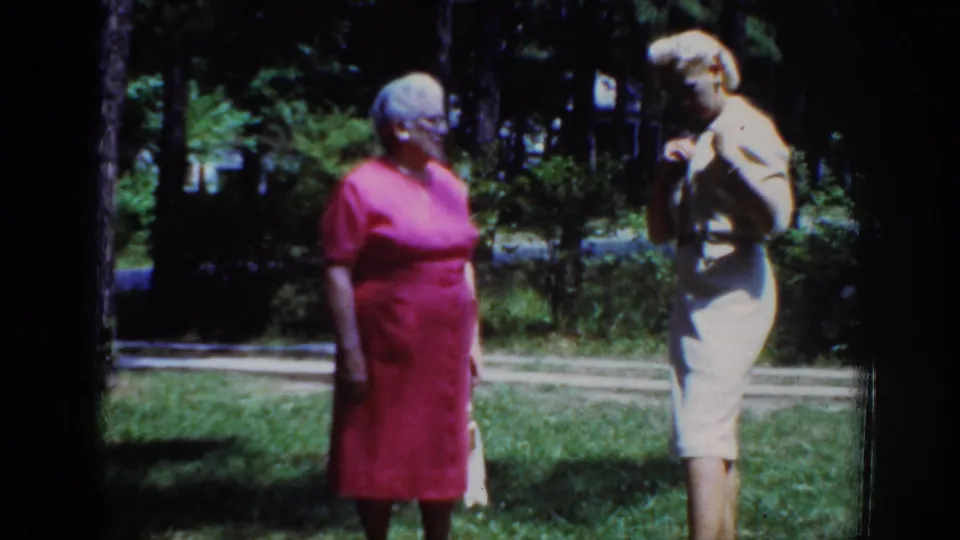 1962 Raleigh home-movie frame showing two adult women conversing outdoors, with grass, trees, and strong sunlight behind them.