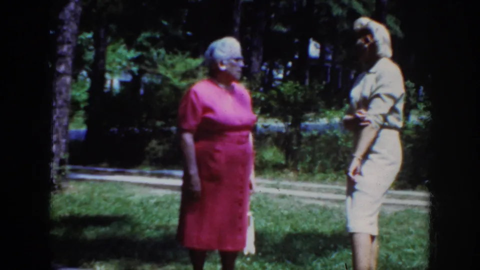 1962 Raleigh home-movie still showing a woman in a pink dress and glasses facing a blonde woman in a light dress outdoors.