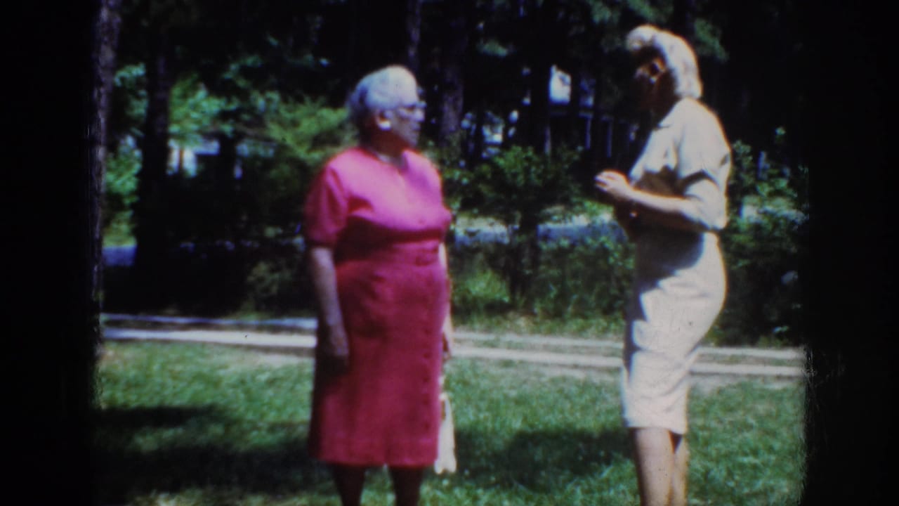 In 1962 Raleigh home-movie footage, two adult women in pink and light dresses face each other on a shaded lawn.