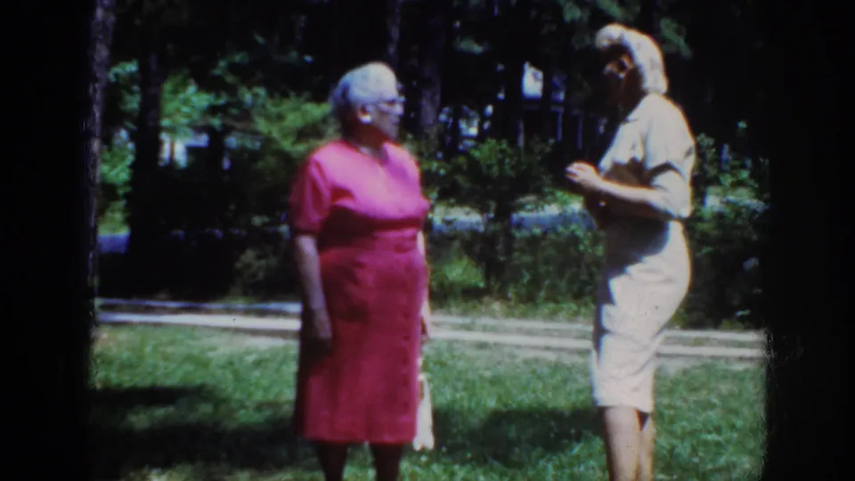 In 1962 Raleigh home-movie footage, two adult women in pink and light dresses face each other on a shaded lawn.