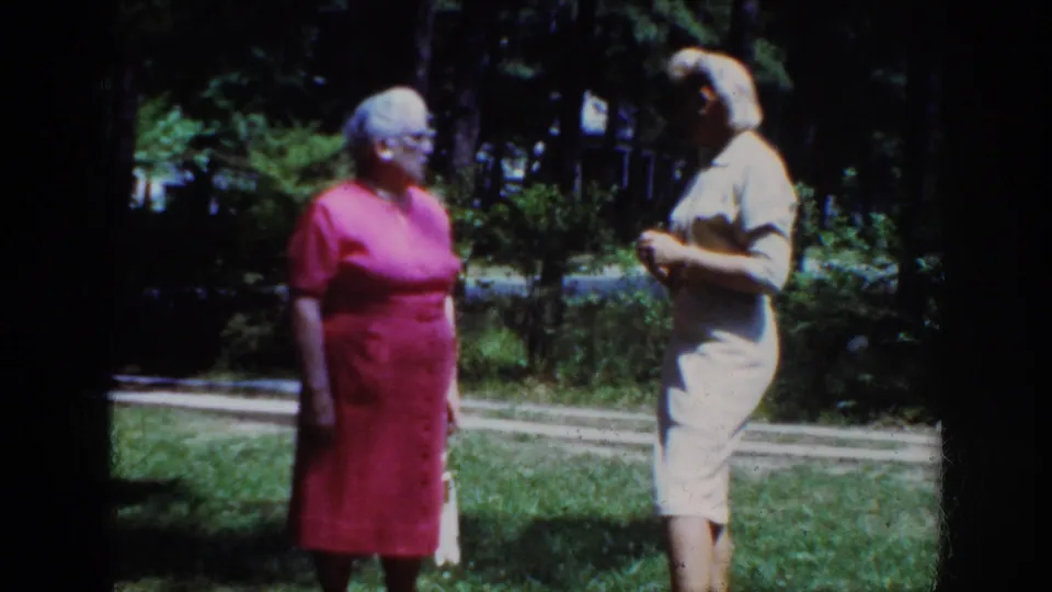 The women continue their outdoor conversation in Raleigh, with the woman in pink looking toward the woman in the light dress.