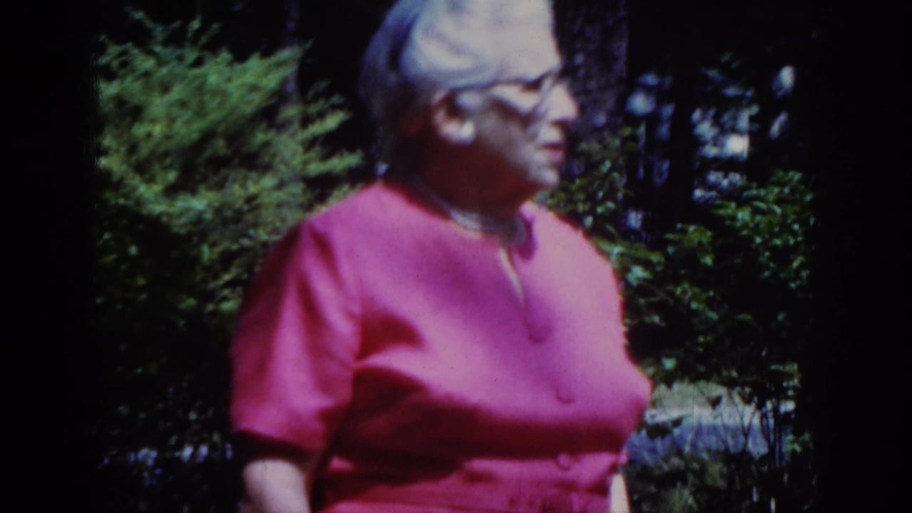 Older woman in a bright pink dress and glasses stands in a wooded Raleigh outdoor setting in 1962 home-movie footage.