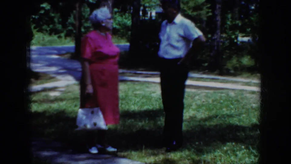 The older woman in pink with a white handbag stands near a man on a grassy wooded area in Raleigh 1962 footage.