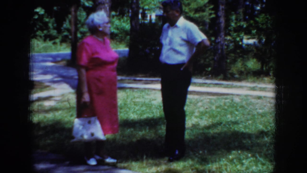 In metadata-supported 1962 Raleigh, an elderly woman with a white handbag stands facing an elderly man beside grass and trees.