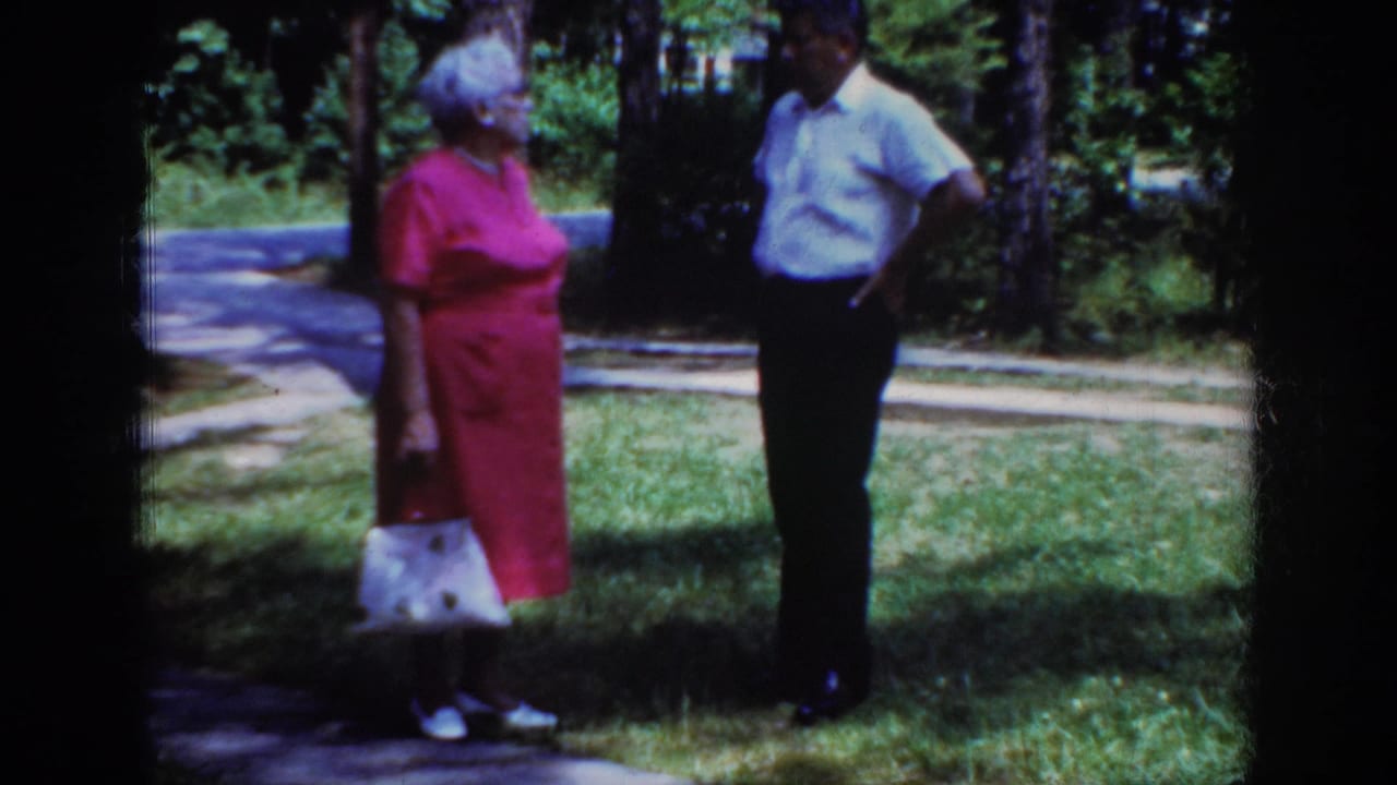 Elderly couple stands near a path with trees behind them.