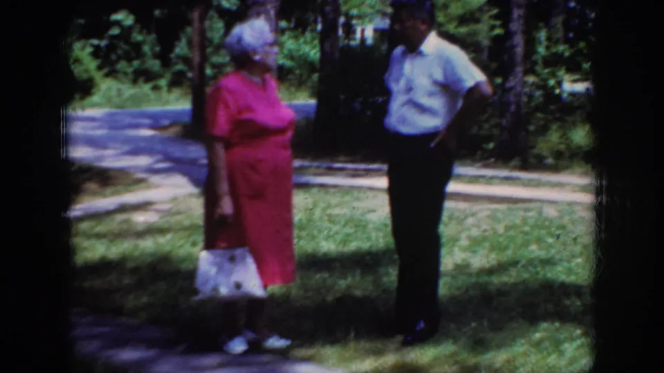 A soft 8mm frame shows the elderly couple standing on a grassy Raleigh lawn, with trees and a curving path in the background.